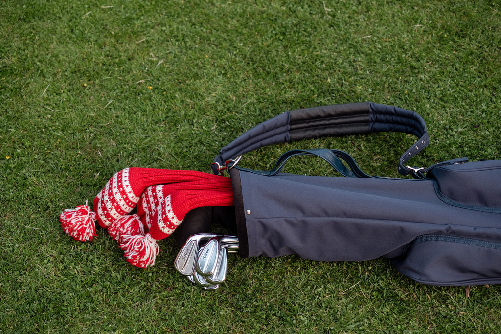 Red wool golf headcovers, knitted with inspiration from Nordic patterns, shown in a classic carry bag on green grass – premium golf accessories for discerning players who appreciate heritage and style.